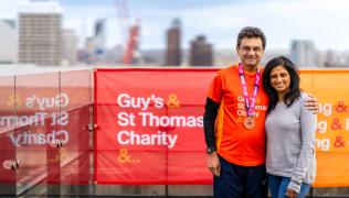 Sunil with his wife, Nisha, stood on a roof terrace with the backdrop of London, after running the 2024 London Marathon. Sunil is wearing a Guy's & St Thomas' Charity red top and is stood next to a red and orange banner that reads 'Guy's & St Thomas' Charity.' 
