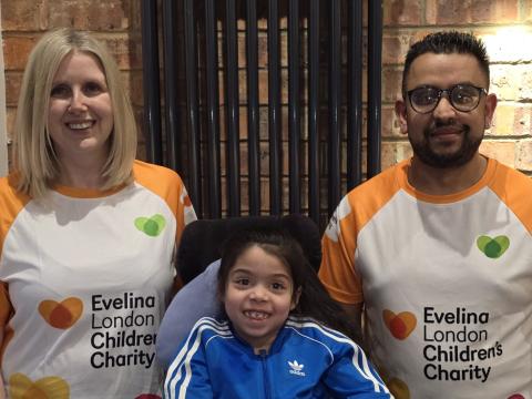 Sarah and Danny stood either side of Sophia who is sat in her wheelchair. They are all smiling. Sarah and Danny are wearing orange and white Evelina London Children's Charity marathon t-shirts, and Sophia is wearing a blue tracksuit top with white stripes. 