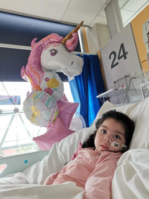 Sophia in Evelina London Children's Hospital on her 5th birthday. She is wearing a pink top and she is in a hospital bed, behind her are birthday balloons. 
