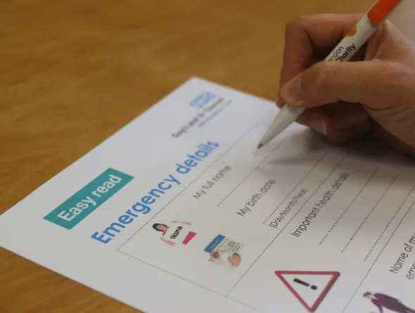 Alt text A hand holding a pan is completing a paper form. The form says 'Easy read' at the top of the page, and contains blank spaces to complete about a patient's emergency details.