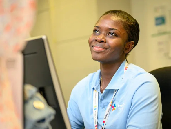 A patient access officer sits behind a computer, looking up and smiling warmly at a visitor