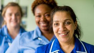 3 female and ethnically diverse nurses wearing blue uniform smile at the camera