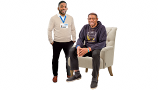 A Black male health professional in his own clothes but with an NHS ID stands next to a Black man sitting in an armchair. They are both smiling
