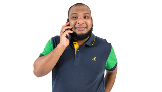 A Black man with a beard dressed in a casual T-shirt holds a mobile to his ear and smiles