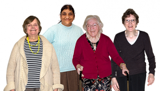 4 older women stand together smiling, one using a walking aid