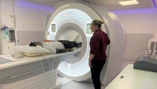 A radiographer in a maroon t-shirt is standing by a Siemens MRI machine. A patient is on the MRI scaner table.