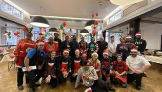 A team photo of catering staff at St Thomas' Hospital. There are about 20 of them, in uniform with Christmas jumpers, hats or aprons on. They are standing or kneeling in a large group.