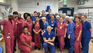 A group photo of the surgical and theatre teams who did the Aquablation HIT list. They are wearing maroon and blue scrubs and are standing or kneeling and smiling.