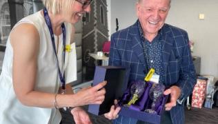 Sue Cox, Associate Chief Nurse, hands Mario a box with two crystal goblets. They are both smiling Sue is wearing a white sleeveless top, Mario a blue checked smart jacket and blue top.