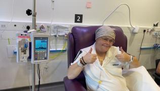 Claire is sat in a purple chair, smiling, with her thumbs up. She is receiving chemotherapy treatment from a machine to the left of her chair. 