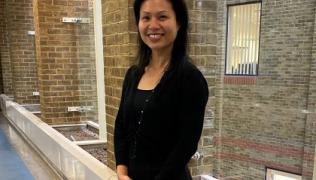 Dr Sui Wong is standing in a corridor, with a brick wall behind her. She is wearing  a black top and cardigan, and has her hands clasped in front of her. She is smiling.