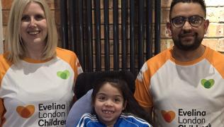 Sarah and Danny stood either side of Sophia who is sat in her wheelchair. They are all smiling. Sarah and Danny are wearing orange and white Evelina London Children's Charity marathon t-shirts, and Sophia is wearing a blue tracksuit top with white stripes. 