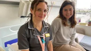 Nurse wearing a dark uniform with a name badge sits beside a woman in casual clothes on a hospital bed near a window overlooking London.