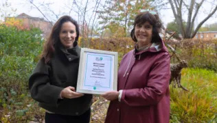 Two people standing in a garden holding a certificate. The person on the left is wearing a black jacket and the person on the right is wearing a red jacket.