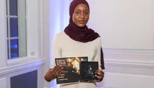 A woman wearing a white tunic and a burgundy headscarf is holding a certificate marked 'Rising Star' and an engraved object.