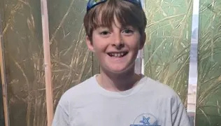 James is wearing a white t-shirt with a blue logo on his left side. He has sunglasses on his head and has light brown, tousled hair. He is smiling into the camera.