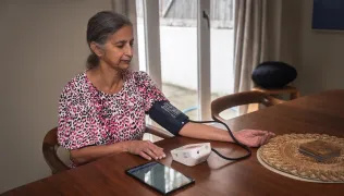 A woman sits at a table at home and takes a blood pressure reading