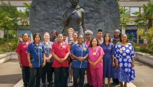 A large group of healthcare professionals and staff stand in front of a large bronze statue. Wreaths and flowers are in front of them.