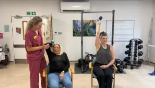 A physio, in maroon scrubs, is helping a seated patient to lift a dumbell. Another patient is seated nearby, and is lifting a dumbell above her head with her right arm.