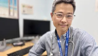 Dr Ronny Cheung, consultant paediatrician at Evelina London, sits turned away from his desk and computer and has a warm expression. He wears a grey shirt, blue lanyard and a stethoscope