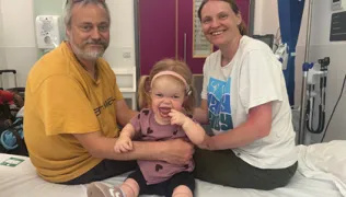 Maisie is sitting on a hospital bed with her parents Ian and Michelle 