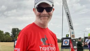 Dave is wearing a red t-shirt with Harefield written in white on the front. He is holding an archery bow, and is standing in front of targets.