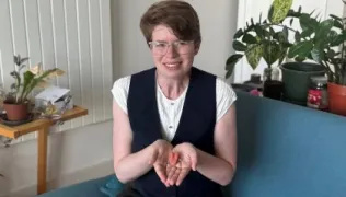 Imogen, wearing a white top and navy waistcoat, is sitting on a sofa, holding her prosthetic tongue in her hands in front of her.