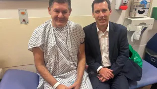Patient Nicholas Kokkinos and surgeon Professor James Gossage are pictured sitting next to each other on a blue clinical bed. Nicholas is wearing a hospital gown and James is wearing a white shirt and dark suit.