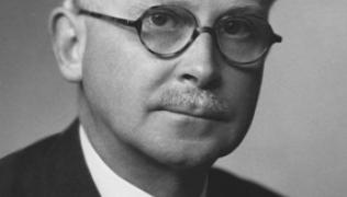 A black and white photograph of Harold Ridley. He is a white man with short white hair and glasses. He is wearing a dark suit