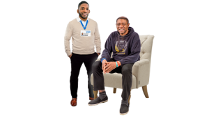 A Black male health professional in his own clothes but with an NHS ID stands next to a Black man sitting in an armchair. They are both smiling