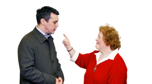 A smartly dressed white woman points a finger at a smartly dressed white man who holds his hands together