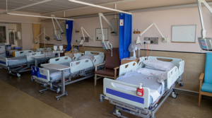 Empty ward with a row of 3 hospital beds separated by blue curtains. Each has a bedside table, chair for visitors and television screen