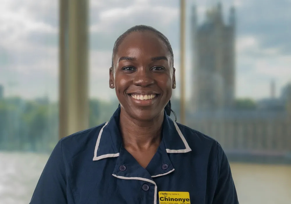 Midwife Chinonye Iroegbulem smiles at the camera. Behind her, out of focus, you can see views of the river Thames through a window in the home from home birth centre at St Thomas' Hospital.