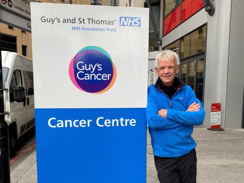 A photo of Geoff May stood next to a sign outside the Cancer Centre at Guy's. Geoff is an older white man with short grey hair and is wearing a blue jacket with his arms folded, looking at the camera.
