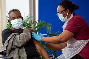 Patient wearing a face mask and a black t-shirt, receiving his COVID-19 vaccination by a nurse wearing a face mask, blue surgical gloves and a white plastic apron.