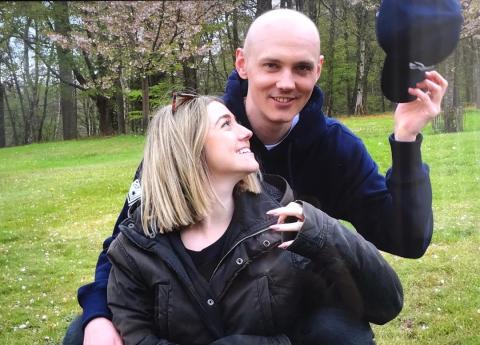 A photo of Troy Randall stood behind his fiancé Sophie. Troy is a white man in his late 20s/early 30s. He has a bald head and is holding a cap in his hand. Sophie is a young white woman with blonde shoulder length hair. She is looking up at Troy.