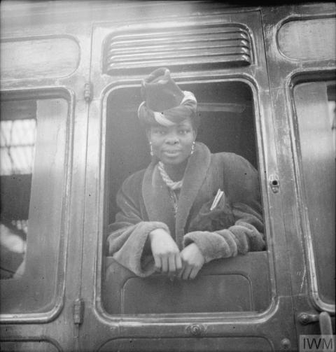 A black and white photograph of Princess Omo-Oba Adenrele Ademola looking out the door of a train. She wears a hat and fur coat.