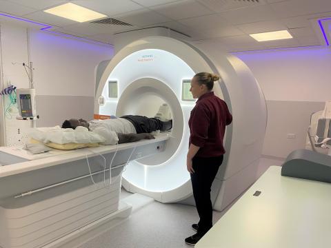 A radiographer wearing a maroon t-shirt and trousers is standing by an MRI scanner. A patient is on the scanner table.