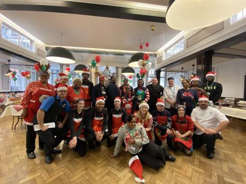 A large group photo of some of the catering team at St Thomas' Hospital. They are wearing their uniforms, with Christmas hats, aprons or jumpers and are standing or kneeling in Shepherd Hall.