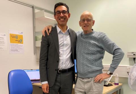 Professor Bijan Modarai is standing in a clinic room, wearing a dark suit and shirt. Patient Kevin Dowd is next to him, with his arm round his shoulder. He is wearing a grey jumper and trousers.They are both smiling.