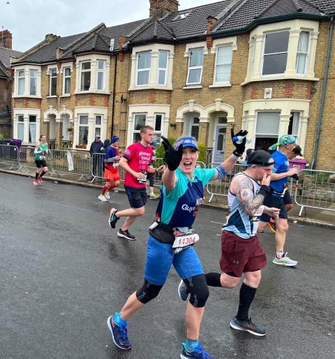 Naomi Ashley-Thorne running in the London Marathon. She wears a blue cap, blue t-shirt and cropped blue leggings. In the background there are runners, spectators and houses.  