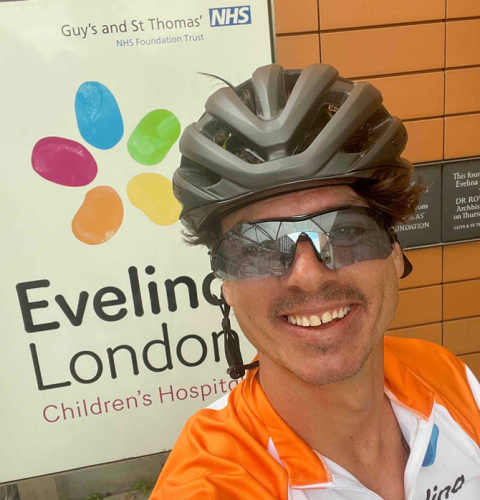 Scott by the Evelina London sign during his challenge. He is smiling and wearing an Evelina top.