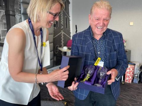 Sue Cox, Associate Chief Nurse, hands Mario a box with two crystal goblets. They are both smiling Sue is wearing a white sleeveless top, Mario a blue checked smart jacket and blue top.