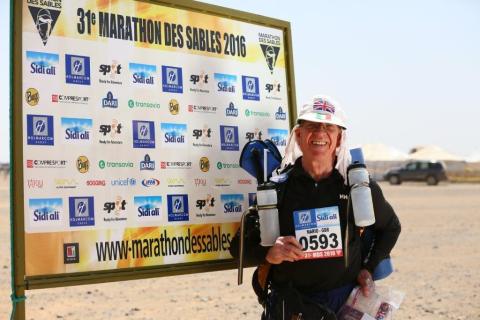 Mario is wearing running gear and has water bottles. He is standing next to a signboard in the desert. It says 31 Marathon des Sables 2016.