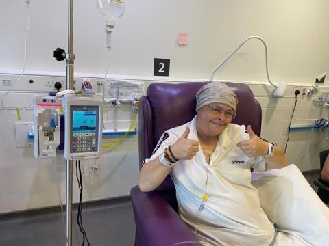Claire is sat in a purple chair, smiling, with her thumbs up. She is receiving chemotherapy treatment from a machine to the left of her chair. 