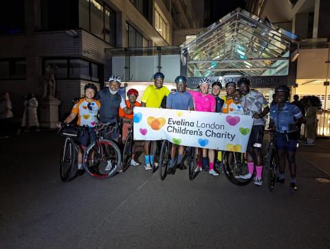 Cycling team outside St Thomas' Hospital at night. The team are holding an Evelina London Children's Charity banner. 