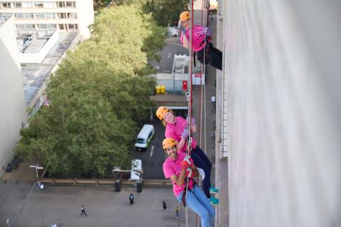 3 people in pink t shirts and orange helmets smiling as they abseil down St Thomas' hospital building. 