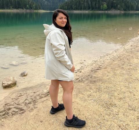 Aila Collins is wearing a beige rain jacket, and is standing in front of a lake. She has long, dark hair.