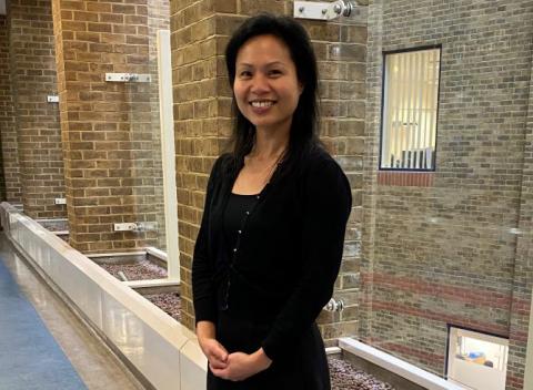 Dr Sui Wong is standing in a corridor, with a brick wall behind her. She is wearing  a black top and cardigan, and has her hands clasped in front of her. She is smiling.