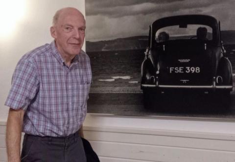 Brian is standing on the left of the image, wearing a checked short-sleeve shirt and dark trousers. To his right is a photo of a vintage car.